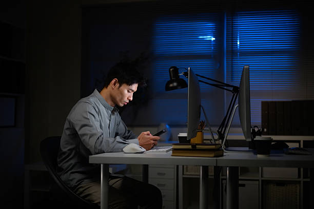 istock Young businessman working late at night in office, focused on smartphone beside computer 2233105681