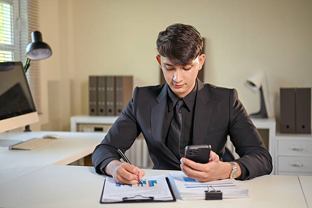 istock Young businessman analyzing financial reports while checking smartphone at office desk 2233105747