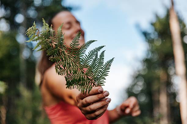 A woman in her 20s holding a fern leaf in a forest setting, with a blurred background of trees and sky.
