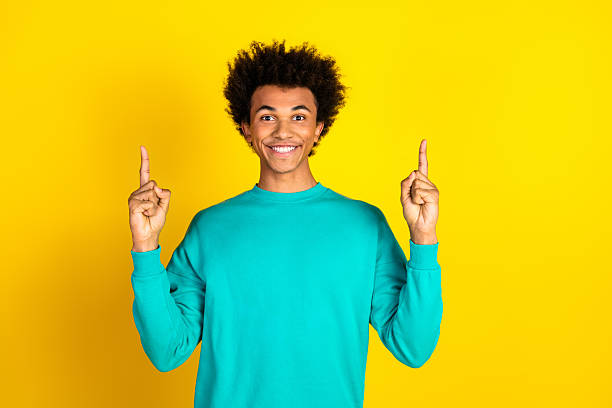 istock Smiling young man in teal sweatshirt gesturing upward with both hands against a vibrant yellow background 2233125926