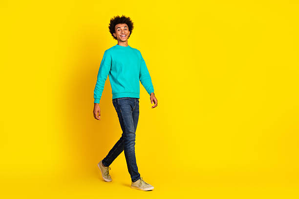 istock Young smiling man in teal sweatshirt walking confidently against a bright yellow background, showcasing casual style and happiness 2233126029