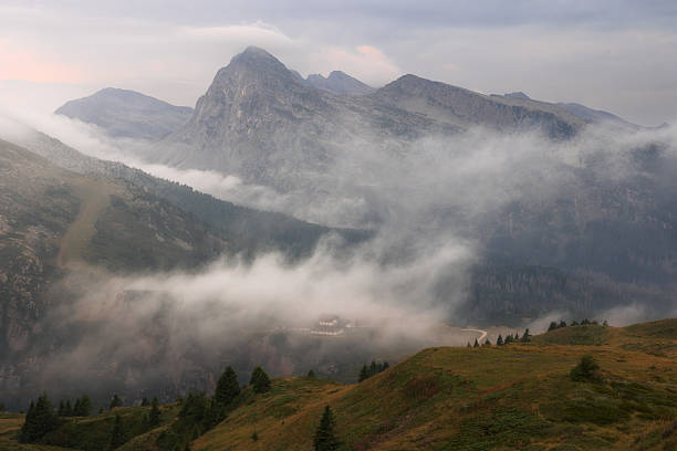 A solitary refuge emerges from drifting mists, sheltered beneath towering peaks. The mountains stand timeless and still, as clouds weave their silent veil across the valley, blurring the line between reality and dream.
