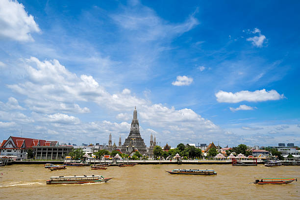 Bangkok, Thailand – August 6, 2024\nEditorial image showing Wat Arun, the Temple of Dawn, one of Bangkok’s most iconic landmarks. Located on the west bank of the Chao Phraya River, the temple features a central prang (tower) surrounded by four smaller ones, all intricately decorated with colorful porcelain. In the foreground, boats are seen navigating the river, adding a dynamic element to the serene architectural scene. The photo captures the cultural and spiritual essence of Thailand’s capital under a clear sky.
