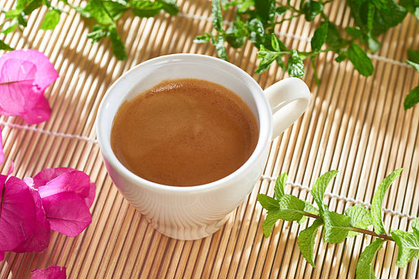 Cup of fresh espresso sits on a bamboo mat, adorned with sprigs of mint and bougainvillea blossoms, creating a tranquil and inviting scene