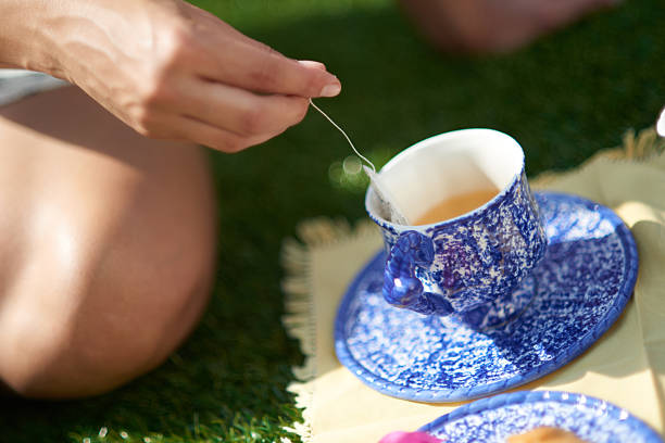 Woman dipping a tea bag in a cup of hot water, sitting on grass during a picnic on a sunny day
