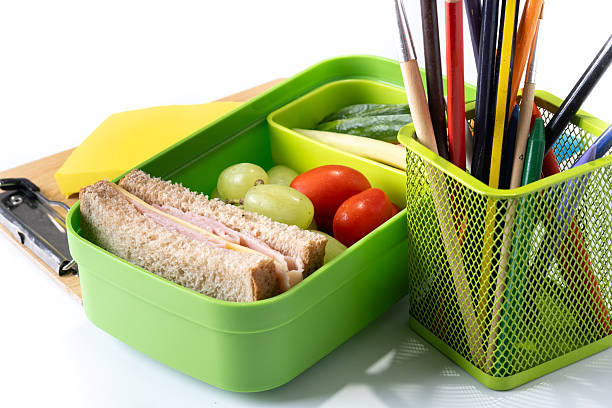 A table top view of green hardback book lies beside a metal lunch box containing a sandwich, set on a wooden surface for a school or study scene. Back to school