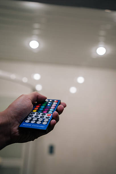 Close-up hand holding RGB remote control, poised to adjust smart lighting system, changing ambiance of room or setting specific mood with colorful led lights.