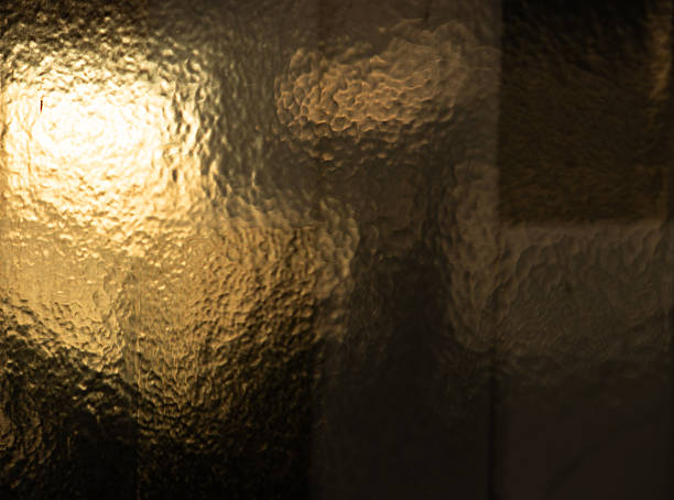 A beautiful abstract background featuring the texture of hammered or patterned privacy glass, illuminated by a warm, golden light. The play of light and shadow creates a sophisticated, elegant, and vintage atmosphere, perfect for luxury design projects or as a subtle backdrop.