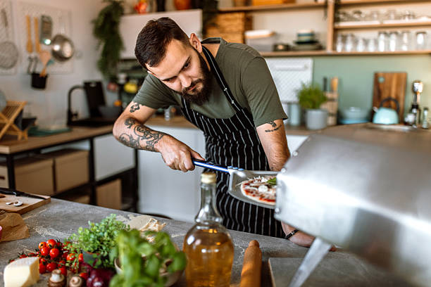 istock Young man in kitchen launching pizza in oven, using pizza shovel. 2233177094