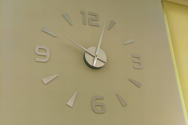 Modern wall clock with silver hands and oversized numbers on a beige wall, minimal design and soft light
