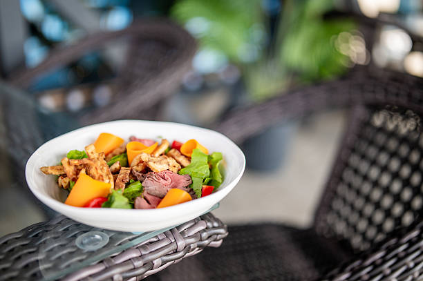 Fresh salad with roast beef, cheese, bell pepper, and crispy toppings in a white bowl on a rattan chair, outdoor dining setting with natural light and relaxed atmosphere