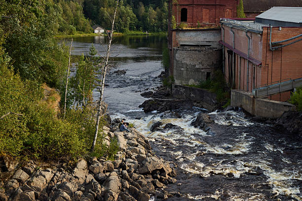 A tranquil riverscape features gentle flowing water located next to decaying and abandoned industrial structures