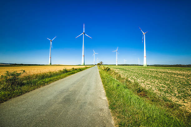 Wind turbines in a vast rural field bordering a straight paved road with blue skies representing renewable energy and sustainable practices.