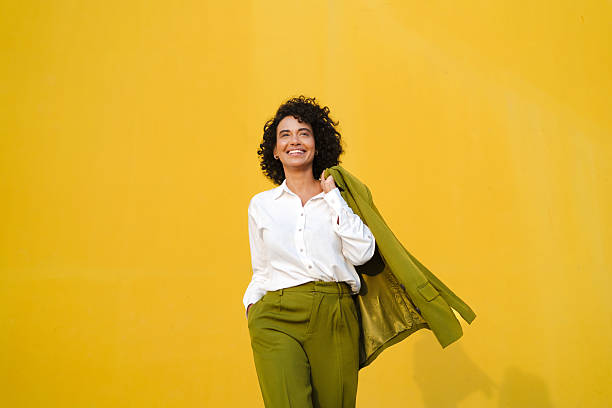 istock Confident businesswoman posing with jacket draped over shoulder, smiling against bright yellow background, embodying professional success 2233227992