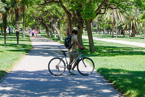 Valencia, Spain - July 13, 2025: Young man walking with bicycle along a lush green path in a park, surrounded by trees and other visitors enjoying the sunny day.