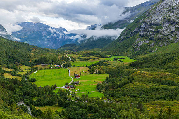 Vibrant valley scene featuring rolling green fields, charming farmhouses, and majestic mountains partially covered in clouds, creating a tranquil and picturesque countryside setting.