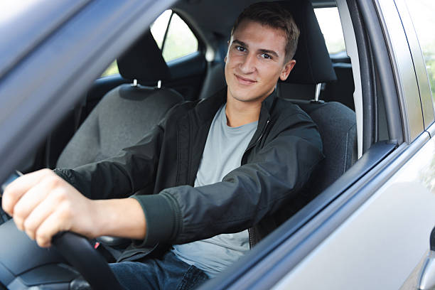 A young man is shown seated in the driver seat of a car, smiling confidently and focusing towards the road, portraying safety, independence, and a cheerful demeanor.