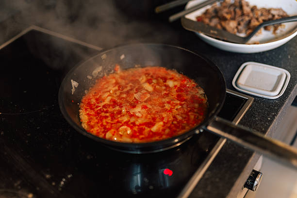 A red tomato-and-onion base simmers in a hot skillet, releasing steam on an electric stovetop. The bubbling mixture forms the aromatic foundation for a hearty home dish of fried plantain with eggs.