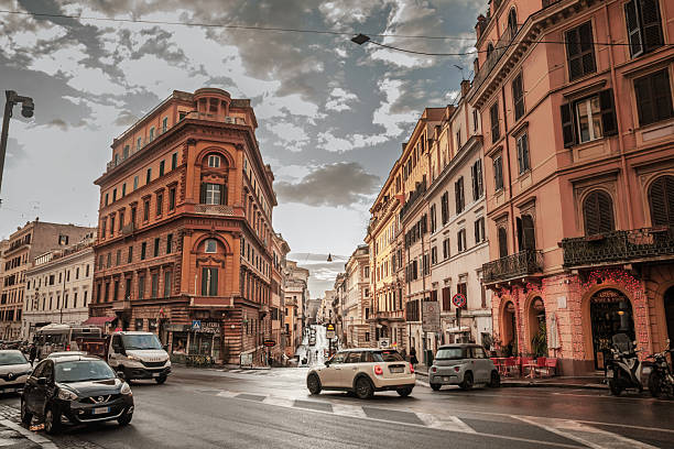 Rome, Italy - January 18, 2025: Urban mobility scene in the Italian capital. Compact cars and scooters navigate a junction lined with ornate facades typical of old Rome. The pavement is damp after rain and headlights catch the reflective surface as vehicles turn across the crosswalk. The architecture frames a narrow avenue that drops toward the horizon, capturing the rhythm of commuting, navigation and tourism in a dense European center. Useful for themes of driving in Rome, traffic behavior, road safety, mobility policy, city logistics, travel features and transport infrastructure.