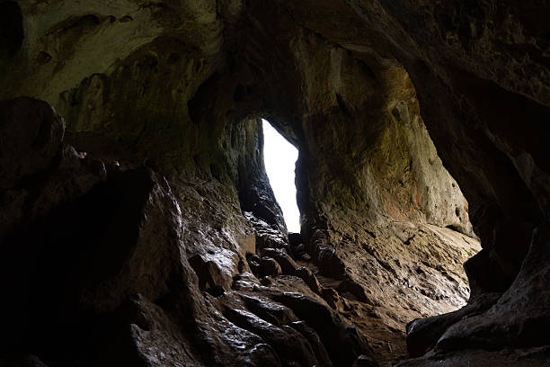 Light streaming through a crack in the ceiling highlights the rough textures of rocks within a dark, mysterious cave, creating enchanting shadows