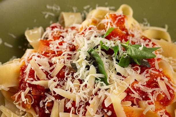 Extreme close-up of a delicious Italian pasta dish. The texture of the grated parmesan cheese, rich tomato sauce, fresh basil, and fettuccine noodles are visible in detail.