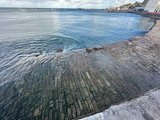 A scenic view of a historic stone slipway gently sloping into calm, blue seawater at high tide. The wet, textured cobblestones glisten in the sunlight as gentle waves lap against the shore. Seagulls fly over the water, and boats are visible on the horizon, adding to the tranquil, coastal atmosphere. Perfect for concepts of seaside charm, maritime history, waterfront tranquility, and coastal architecture.
