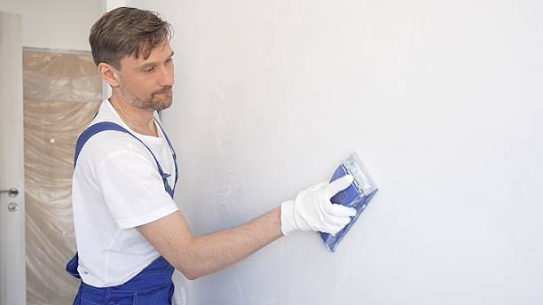 istock Man construction worker wearing protective white gloves and blue construction coveralls, is sanding white wall with blue hand block, preparing smooth surface during interior home renovation project 2233292176