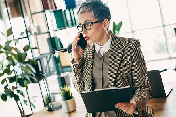 istock Professional mature woman in office calling on phone while reviewing documents, showcasing workplace confidence and style 2233292373