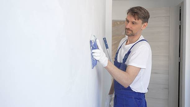 istock Man construction worker wearing protective white gloves and blue construction coveralls, is sanding white wall with blue hand block, preparing smooth surface during interior home renovation project 2233293439