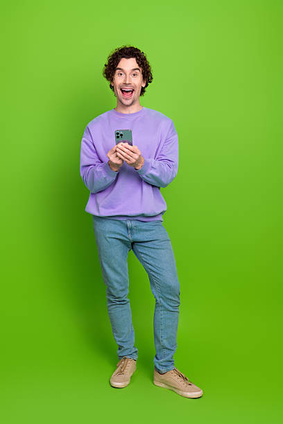 istock Excited young man holding a smartphone against a bright green minimal background, dressed casually in a trendy sweatshirt 2233294517