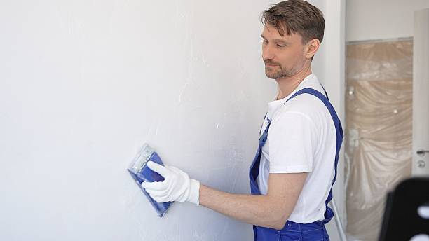 istock Man construction worker wearing protective white gloves and blue construction coveralls, is sanding white wall with blue hand block, preparing smooth surface during interior home renovation project 2233294608