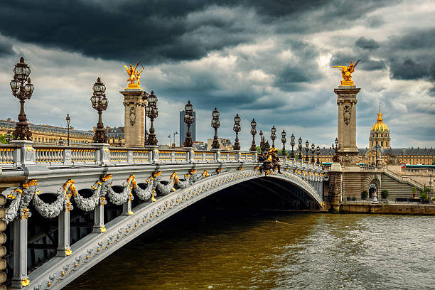 Pont Alexandre III bridge with its gilded sculptures and lampposts, stands under a dramatic and stormy sky over the Seine River in Paris, France.