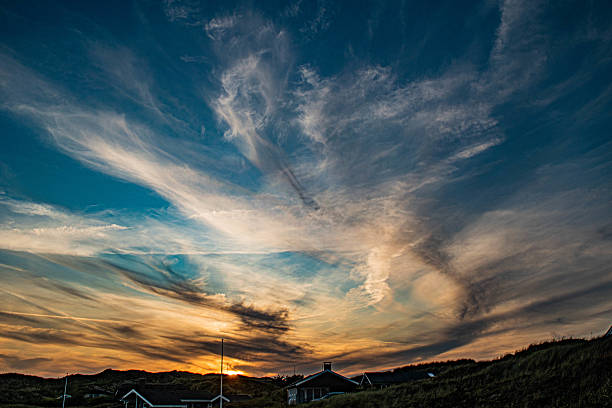 Small houses nestled among sandy dunes with grass on a coastline, under a colorful sunset sky. Warm light and scattered clouds create a peaceful and scenic atmosphere