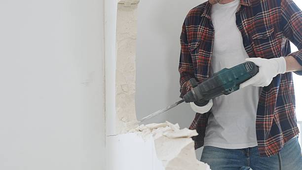 istock Unknown male construction worker wearing red checkered shirt jeans and protective gloves, is demolishing white wall with rotary hammer drill, generating dust, close up view. Renovation concept 2233300267