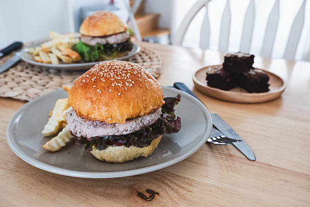 A close-up shot of a gourmet chicken or plant-based burger with sesame seed bun, fresh lettuce, and crinkle-cut fries on a gray plate, with more food in the soft-focused background.