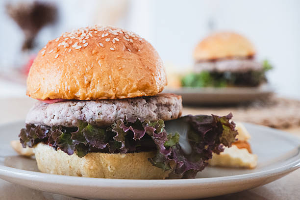 Healthy burger with a light-colored patty and fresh red leaf lettuce on a sesame seed bun, served on a ceramic plate, with another burger blurred in the background.