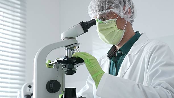 istock Medical scientist wearing protective mask, cap and green gloves using microscope doing scientific research in laboratory, developing vaccine or medication. Medicine, healthcare and science concept 2233302949