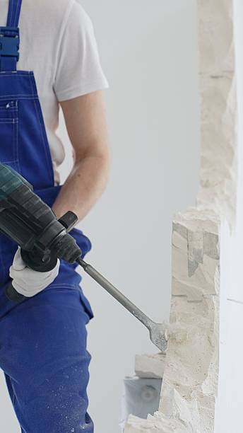 istock Unknown male construction worker wearing blue overalls and protective white gloves, is demolishing white wall with rotary hammer drill, generating dust, close up horizontal view. Renovation 2233303490
