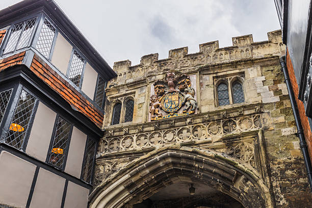 Upward view of Salisbury High Street Gate into Cathedral Close, with carved stonework, battlements, and a colorful coat of arms between timber framed buildings in Wiltshire. Salisbury, UK, 5 July 2024