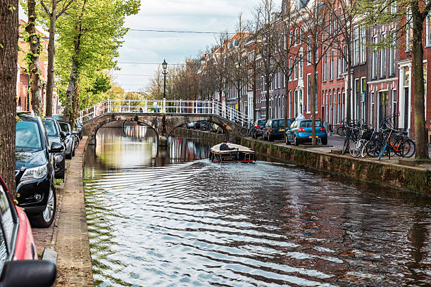 Delft, Netherlands - 26 Apr, 2024 - Scenic Dutch canal with a small boat approaching a historic arched bridge lined with trees and traditional brick houses.