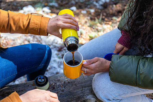 Two hikers are pouring hot coffee from a thermos into a mug while sitting on a log in a winter forest