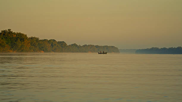 A small boat is gliding on calm waters during sunrise in the Danube Delta, featuring serene reflections and lush greenery along the riverbanks. The scene is peaceful and picturesque, capturing the beauty of nature.