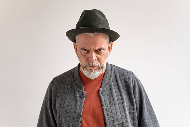 An angry mature Caucasian male is standing against a neutral background, wearing a hat and a casual jacket. He is expressing frustration with a focused and intense gaze.