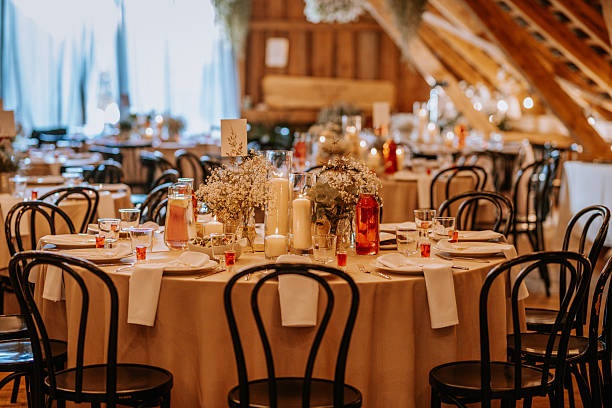 istock Elegant rustic wedding reception setup with round tables, floral centerpieces, candles, glassware, and wooden chairs in a warmly lit venue. 2233319510