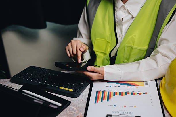 istock Business Professional Analyzing Data on Smartphone in Safety Vest and Hard Hat at Office Desk with Reports and Keyboard 2233320551