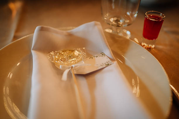 Close-up of a wedding table place setting featuring a white plate, folded napkin, personalized lollipop favor, name card, and a red shot glass for guests.