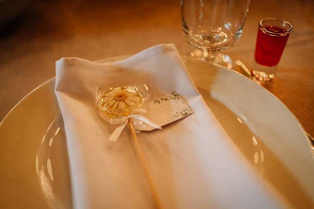 istock Close-up of a wedding table setting featuring a white napkin, elegant plate, floral lollipop with ribbon, name tag, glass, and a red shot glass on a beige tablecloth. 2233320561