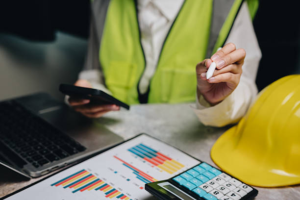 istock Focused Professional Analyzing Data with Calculator and Smartphone While Wearing Safety Vest and Hard Hat in Office Setting 2233320625