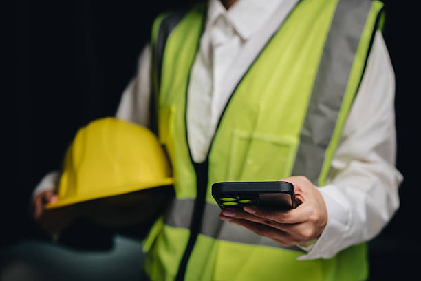 istock Construction Worker in Safety Vest Holding Smartphone and Hard Hat in Indoor Setting with Dark Background 2233321606