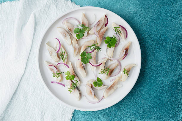 istock Marinated herring slices with red onion and herbs on white plate with napkin over blue background 2233325237